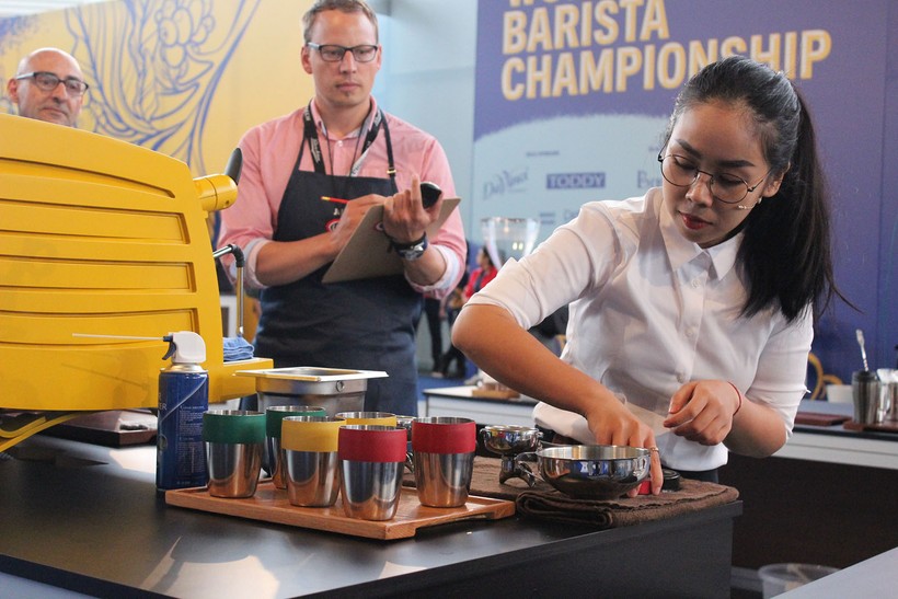 A woman prepares a portafilter to make espresso using a number of tools next to a bright yellow espresso machine. Two men observe as judges, one mostly obscured by the machine and one with clipboard and apron on. In the background are the stage walls, showing a graphic pattern of light and dark blues, accented with yellow and coffee plant motifs